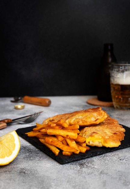 Traditional British fish with chips. On a black plate and on a gray table. Served with lemon and a mug of cold beer. Black background. Space for text