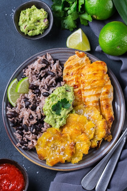 Traditional Central American caribbean cuban colombian food Rice with black beans roasted fried chicken breast and tostones fried green bananas plantains with guacamole sauce Top view flat lay