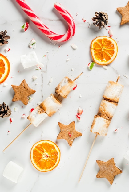 Traditional christmas sweets  candy cane, marshmallow, dried orange, gingerbread stars, baked on fire marshmallow skewers on white marble , copyspace top view