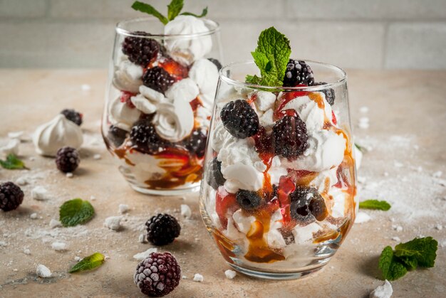 Traditional English dessert. Eton mess  whipped cream, meringue, fresh blackberries, sauce and caramel. In serving glasses on a light stone table. 