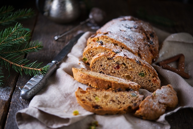 A traditional German Christmas Stollen lies on a wooden table. A celebratory cake is cut into slices with candied fruits, nuts and raisins. Selective focus.