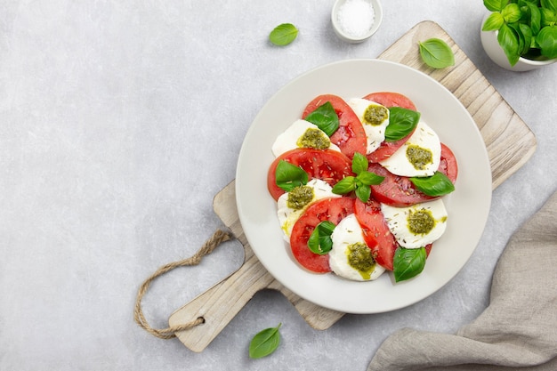 Traditional italian caprese salad with sliced tomatoes, mozzarella, basil, pesto sauce and spice on a light gray concrete background. Top view. Copy space.