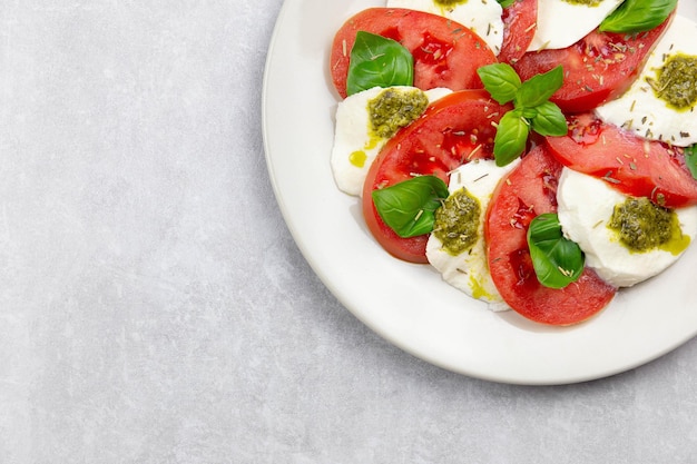 Traditional italian caprese salad with sliced tomatoes, mozzarella, basil, pesto sauce and spice on a light gray concrete background. Top view. Copy space.