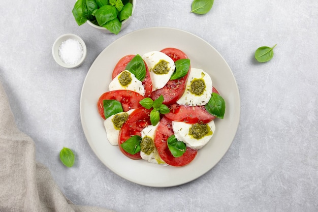 Traditional italian caprese salad with sliced tomatoes, mozzarella, basil, pesto sauce and spice on a light gray concrete surface