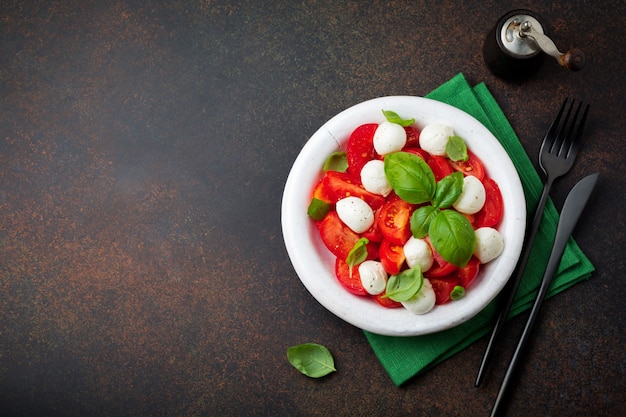 Traditional Italian caprese salad with tomatoes, mozzarella cheese and basil on dark background in white old ceramic plate.