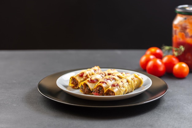 Traditional italian dish cannelloni on a black background in a black plate with tomatoes in the back