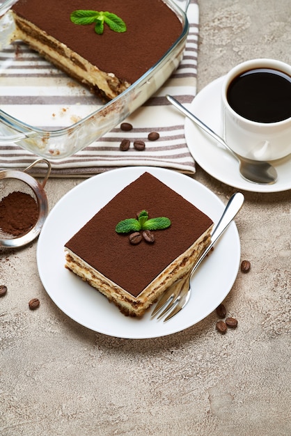 Traditional italian tiramisu dessert in glass baking dish and portion on grey concrete table