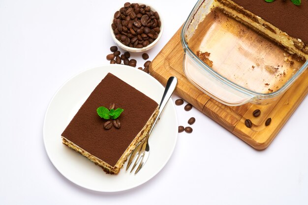 Traditional italian tiramisu dessert in glass baking dish and portion on plate on white