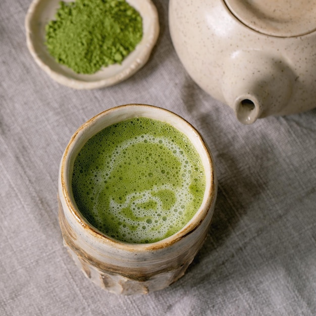 Traditional japanese hot green frothy tea matcha latte in handmade ceramic cup powdered matcha teapot and bamboo whisk standing on grey linen tablecloth Healthy drink Square image