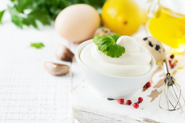 Traditional mayonnaise sauce in white ceramic bowl and ingredients for its preparation on white wooden. 