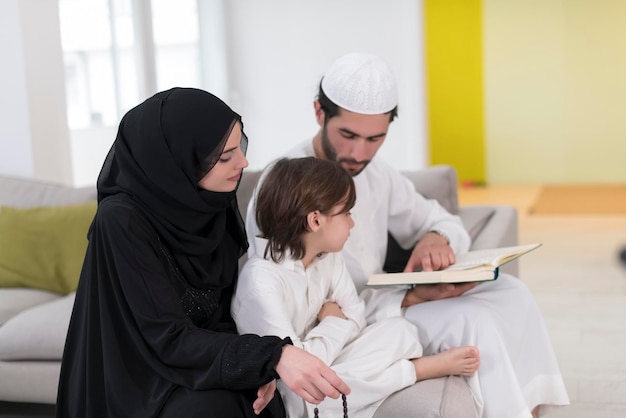 Traditional muslim family parents with children reading Quran and praying together on the sofa before iftar dinner during a ramadan feast at home