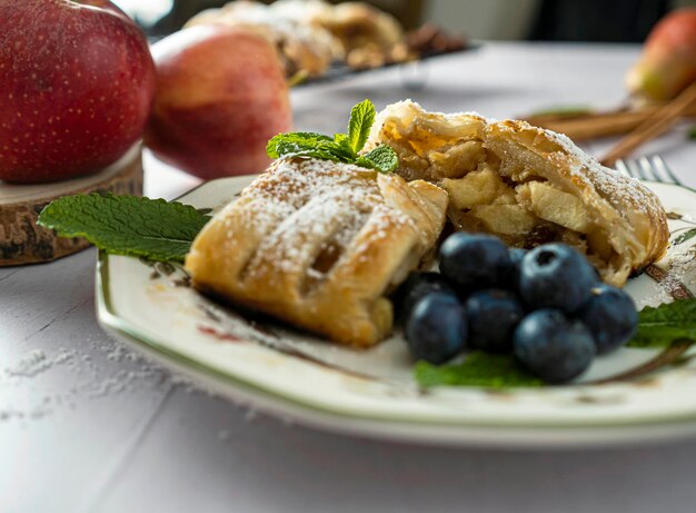 A traditional piece of apple strudel with powdered sugar mint blueberry and cinnamon in the foreground on a table