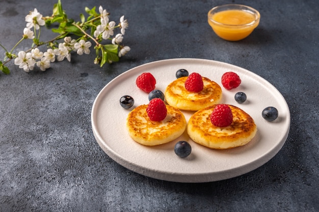 Traditional russian cheesecake with raspberries, blueberries and honey on a white ceramic plate on a dark background