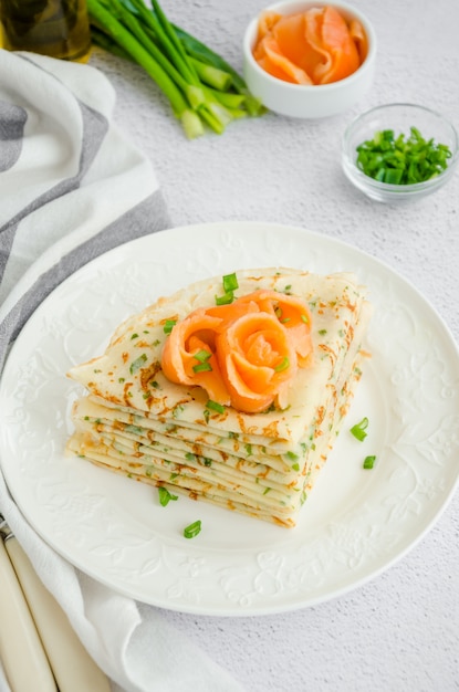 Traditional Russian thin pancakes or crepes with cheese, herbs and smoked salmon on a white plate on a light background. Holiday maslenitsa. Vertical orientation.