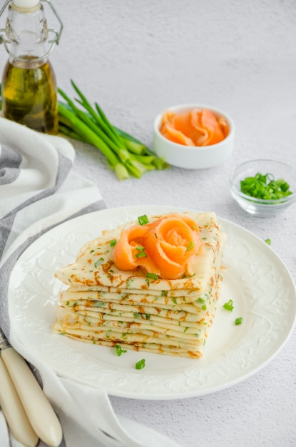 Traditional Russian thin pancakes or crepes with cheese, herbs and smoked salmon on a white plate on a light surface Holiday maslenitsa. Vertical orientation.