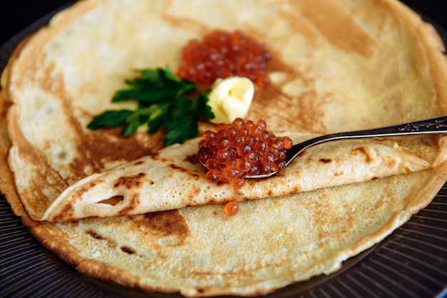 Traditional thin Russian pancakes with red caviar, butter and greens on a black wooden background