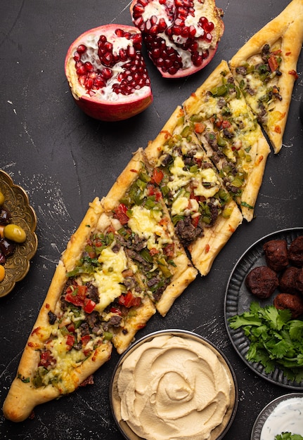 Traditional Turkish pizza pide freshly baked with ground beef and vegetables served on black stone background with assorted Middle Eastern meze from above, cuisine of Turkey