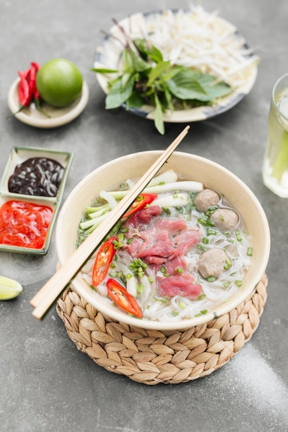 Traditional Vietnamese soup Pho bo with herbs, meat, rice noodles, broth. Pho bo in bowl with chopsticks, spoon. Space for text. Top view. Asian soup Pho bo on wooden table background. Vietnamese soup