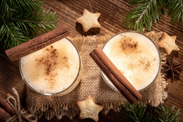 Traditional Xmas eggnog with cinnamon in two glasses fir branches cookies stars on wooden table