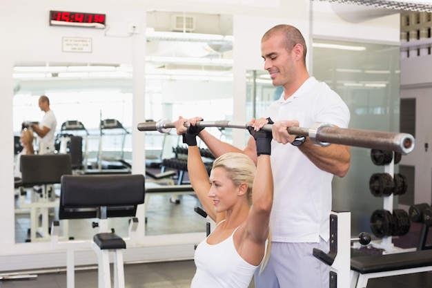 Trainer helping woman with lifting barbell in gym