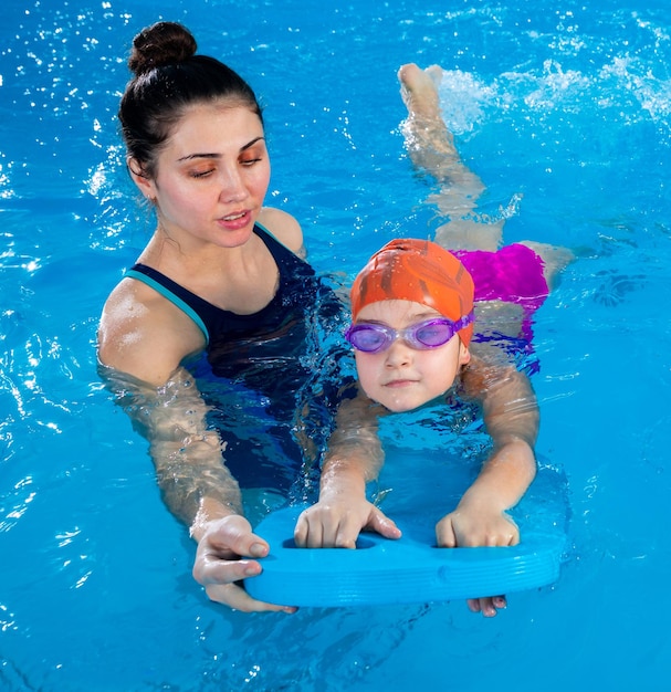 Trainer teaching little girl how to swim in indoor pool with pool floating board