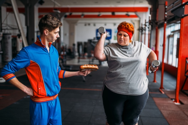 Trainer with hotdog in hand forces fat woman to exercise, motivation, hard workout in gym. Calories burning, obese female person in fitness club, fat-burning, sport against unhealthy food