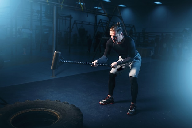 Training in gym, man with sledgehammer hits tire. Active exercise in sport club