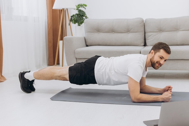 Training At Home Sporty man doing yoga plank while watching online tutorial on laptop exercising in living room free space