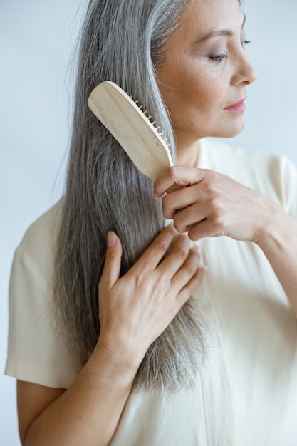 Tranquil middle aged Asian model brushes long hoary hair posing on light background