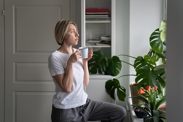 Tranquil middleaged lady drinks hot drink from mug at windowsill looking out of window