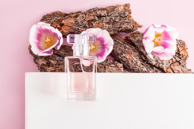 A transparent bottle of women's perfume lies on part of the white podium and bark of a tree with spring flowers presentation of spring fragrance