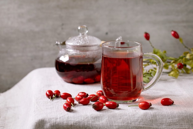 Transparent cup of rose hip berries herbal tea and glass teapot standing on white linen table cloth with wild autumn berries around. Winter hot cozy beverage