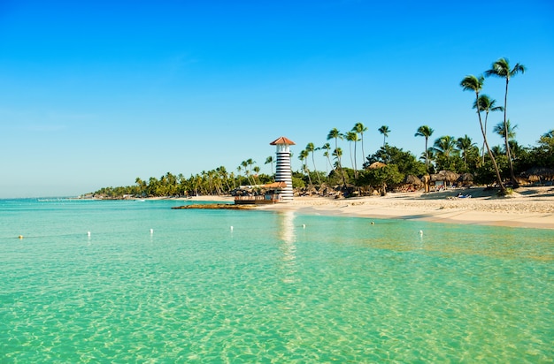 Transparent sea water and clear sky. Lighthouse on a sandy tropical island with palm trees.