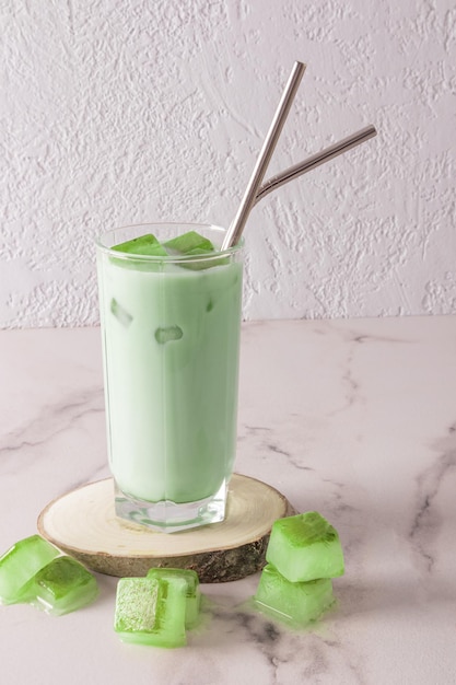 A transparent tall glass with organic cold green matcha tea and ice cubes with ecofriendly metal straw on a wooden natural podium front view