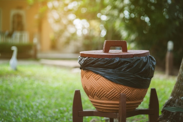 A trash can with a black trash bag sits in a garden.