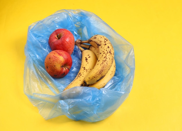 Trash packege with spoiled apples and bananas on yellow background