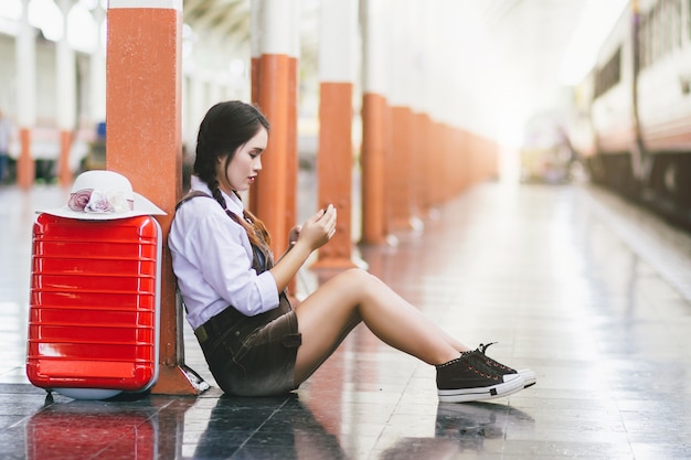 Photo travel asian woman pregnant look at the smartphone with a red suitcase at railway station.