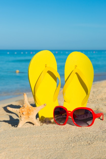 Travel by sea. Beach vacation. Red sunglasses and yellow flip-flops on sandy seashore.