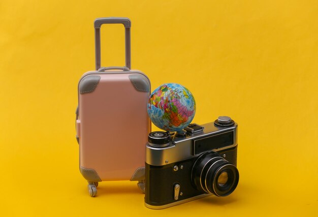 Photo travel concept. mini pink plastic travel suitcase with camera and globe on yellow background. minimal style