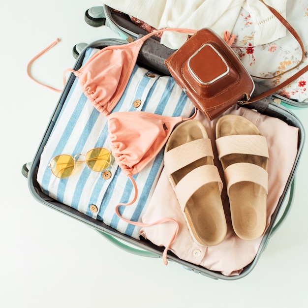 Travel hand luggage with swimsuit bikini, slippers, sunglasses, dress, retro camera on white surface