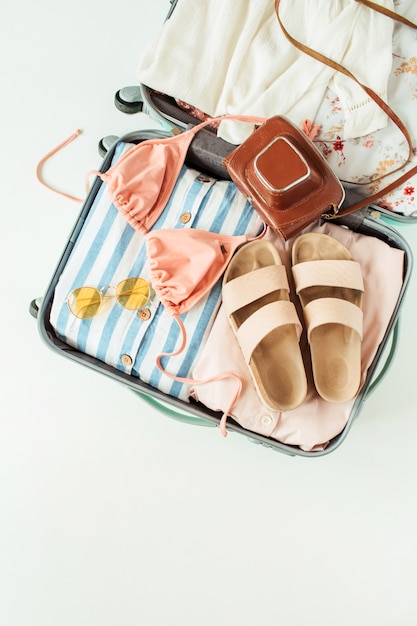 Travel hand luggage with swimsuit bikini, slippers, sunglasses, dress, retro camera on white.