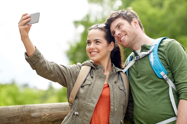 travel, hiking, backpacking, tourism and people concept - smiling couple with backpacks taking selfie by smartphone in nature