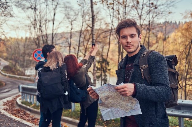 Travel, hitchhiking, adventure concept. Group of young hikers standing on the road sidelines at beautiful autumn forest, guy with map planning hike.