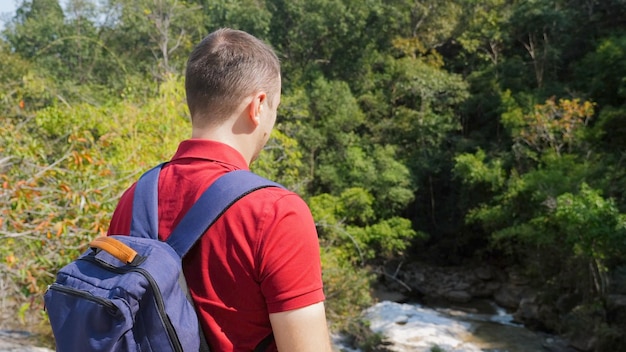 Travel man in red polo shirt with backpack standing near rock river