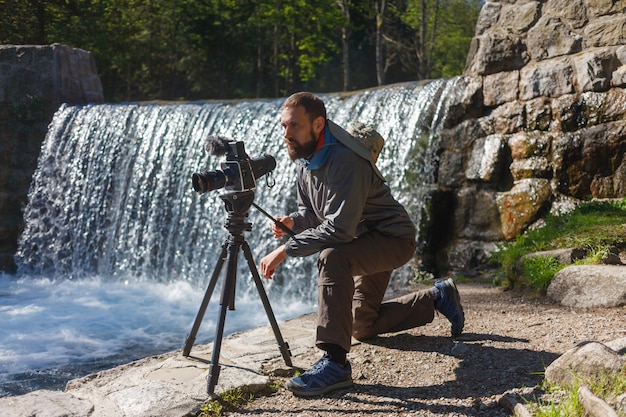 Travel photographer bearded man with professional film camera on tripod shooting mountain landscape in waterfall background. Hiker tourist professional photography shooting, filming backstage