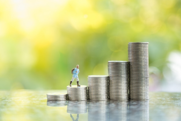 Travel, Saving and planning Concept. Close up of traveler miniature figure people with backpack walking on top of stack of coins with copy space and green nature background.