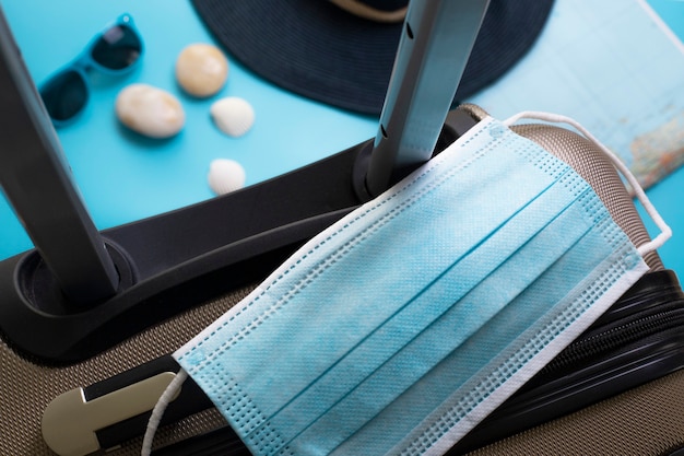 Travel during the time of COVID-19. Medical mask, suitcase, sun glasses, map, seashells on the blue background. Vacation, holidays in corona times. Colorful design.