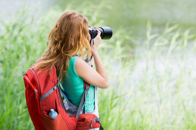 travel, tourism, hike, hobby and people concept - young woman with backpack and camera photographing outdoors