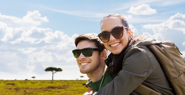 travel, tourism and people concept - happy couple in sunglasses with backpacks traveling over african savannah background