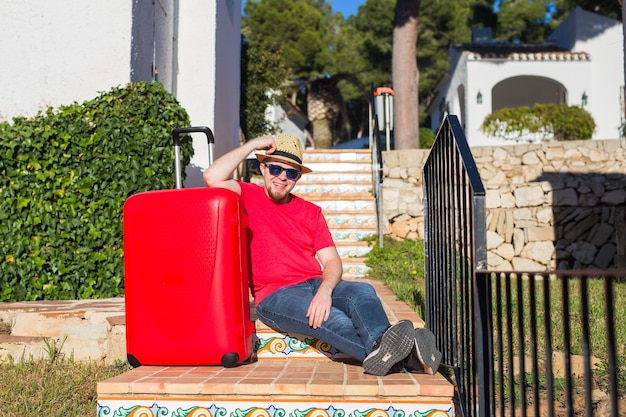 Travel, tourism and people concept. happy man sitting on stairs in a hat with red suitcase.